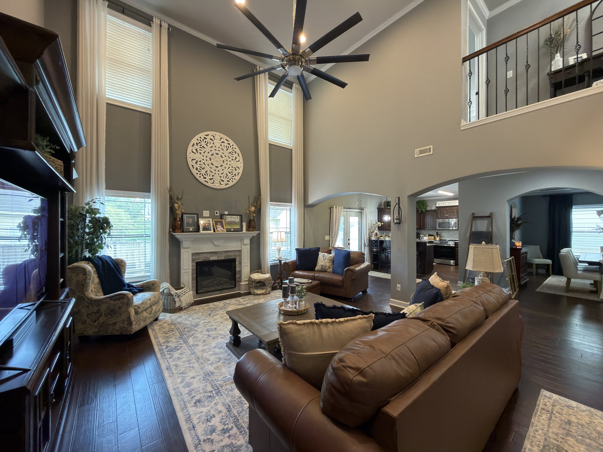Two-story living room with tall windows, a stone fireplace, brown leather sofas and a large ceiling fan, photographed with a phone