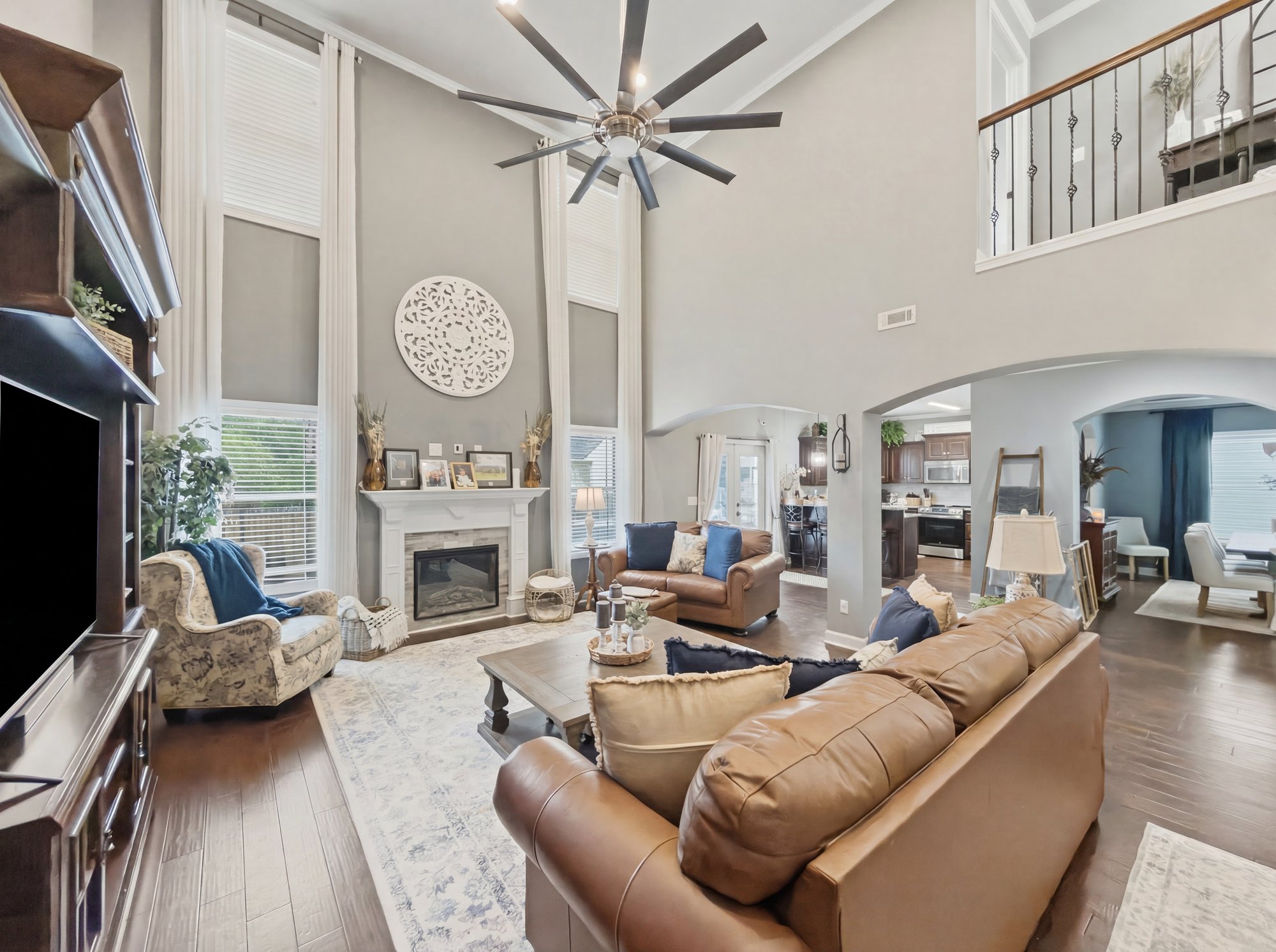 The same two-story living room with tall windows, stone fireplace, brown leather sofas and ceiling fan, professionally edited with brighter, cleaner color