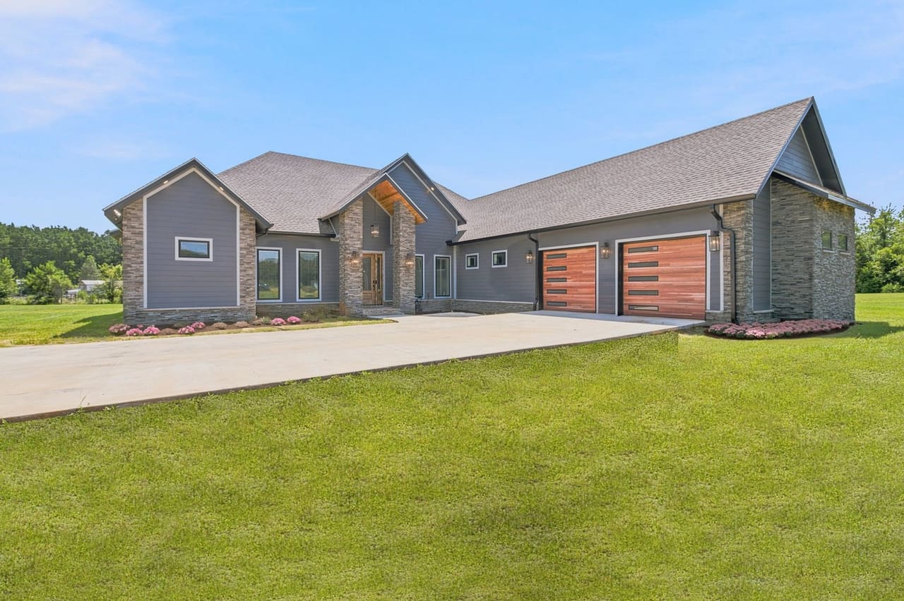 Modern gray farmhouse-style home with stone columns, cedar-toned garage doors and a wide concrete driveway under a blue sky, photographed for a Dothan, Alabama area real estate listing